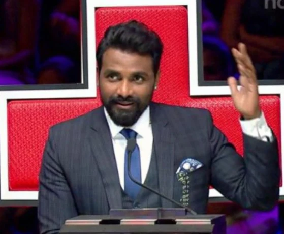 A television dance judge seated at a red judges’ desk.